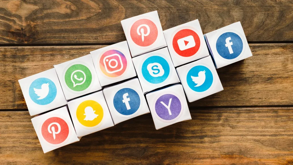 Colorful paper cubes with social media platform logos including Facebook, Twitter, Instagram, YouTube, Pinterest, Snapchat, Skype, WhatsApp, and Yahoo, on a wooden surface.
